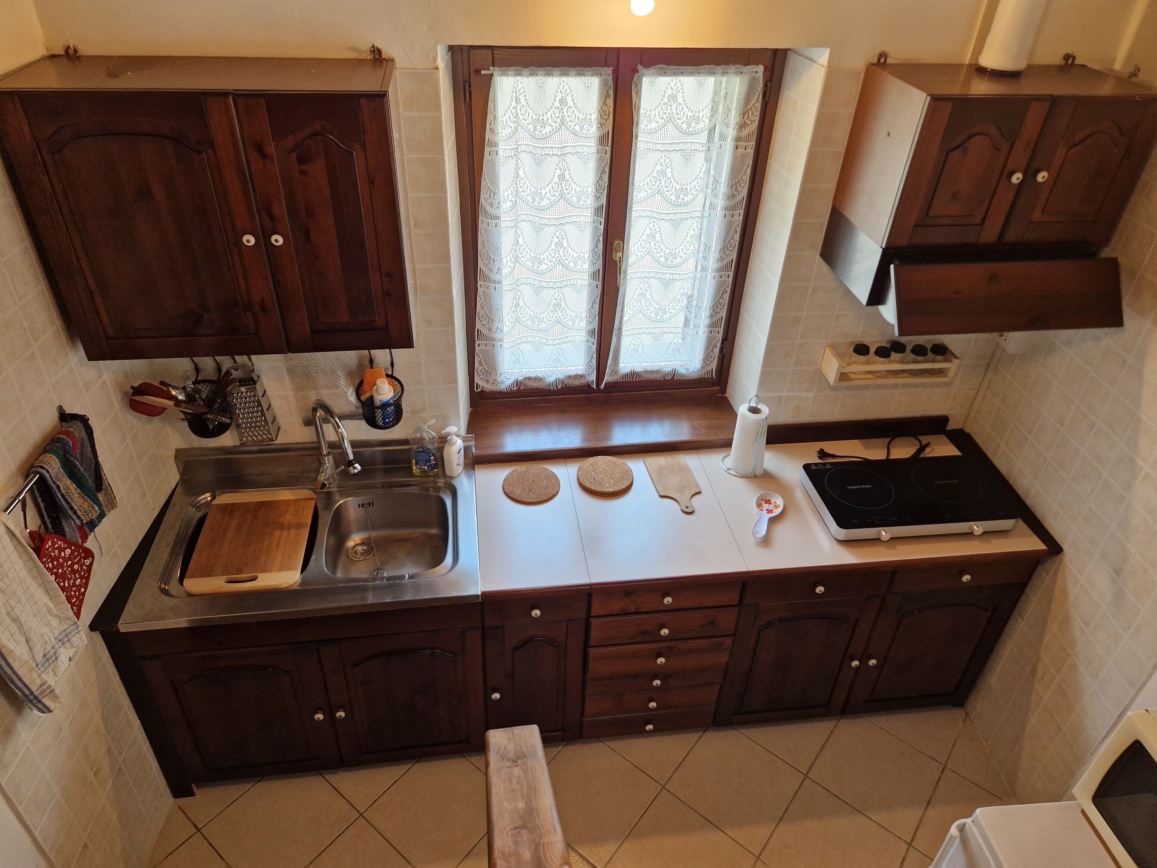Una cocina con armarios de madera oscura, un lavamanos de acero inoxidable, una ventana con cortinas de encaje y una estufa.
