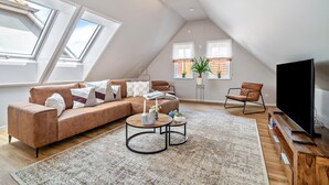 A modern loft apartment with a sloped ceiling, large skylights, a sectional sofa, a round coffee table, a TV on a wooden stand, and a potted plant. - Apartment "Seeterrasse" with Lake View, Shared Terrace and Wi-Fi (Malchow)