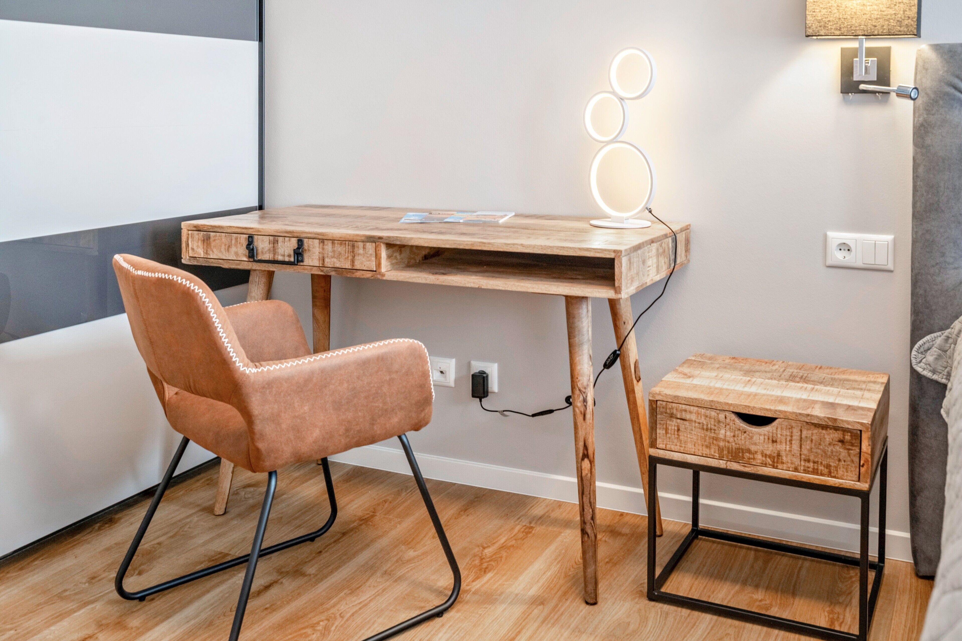 Un bureau en bois avec une chaise, une table de chevet et un luminaire fixé au mur.