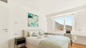 A bedroom with a bed, bedside table, desk, chair, and a window with a view of mountains. - Generoso Luxury Lake View Suite in Lugano (Lugano)