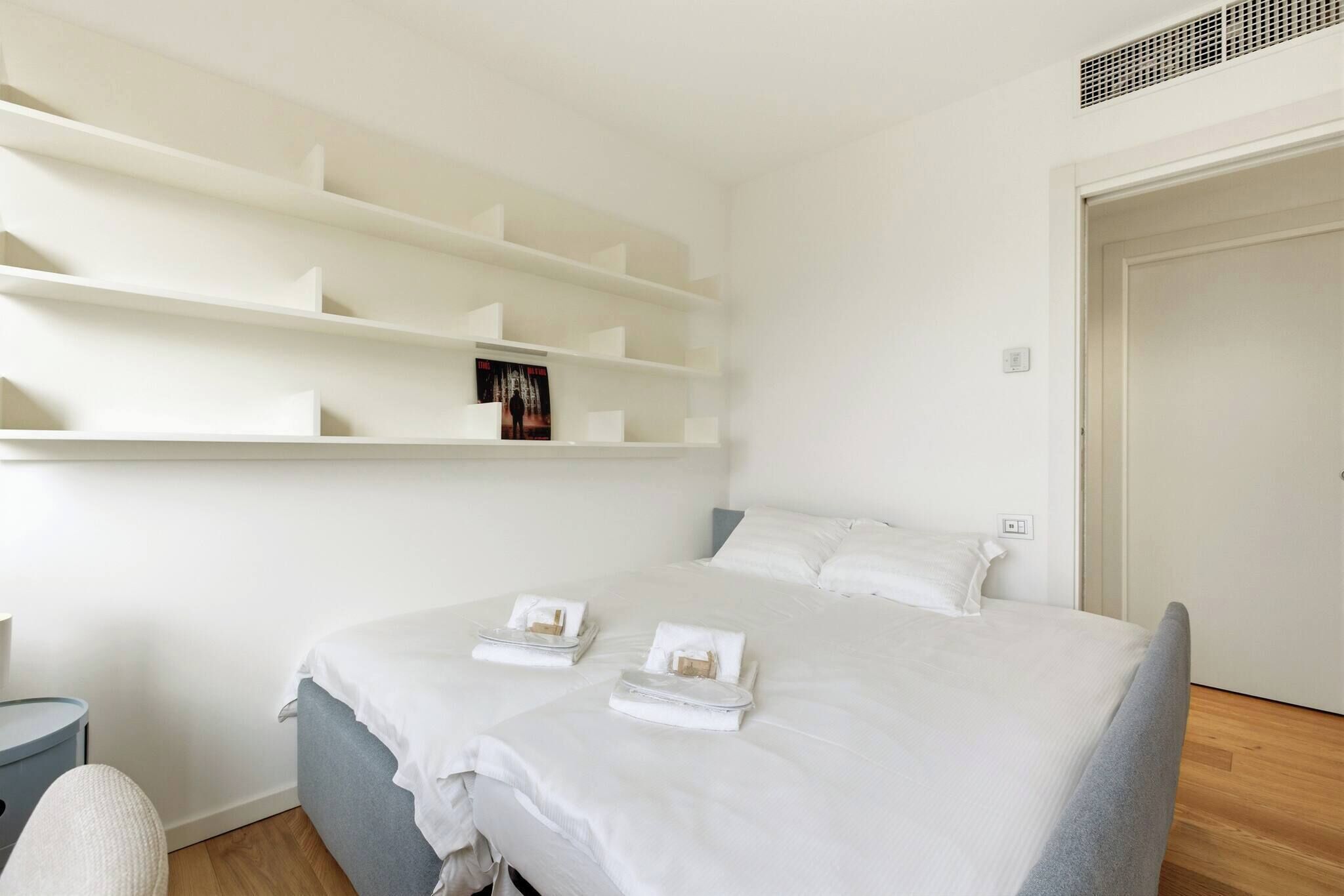 A neatly arranged hotel room with a bed, a bedside table, and a built-in shelf with a book.