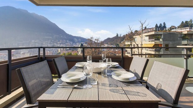 A dining table set with plates, glasses, and cutlery on a balcony with a city and mountain view. - Generoso Luxury Lake View Suite in Lugano (Lugano)