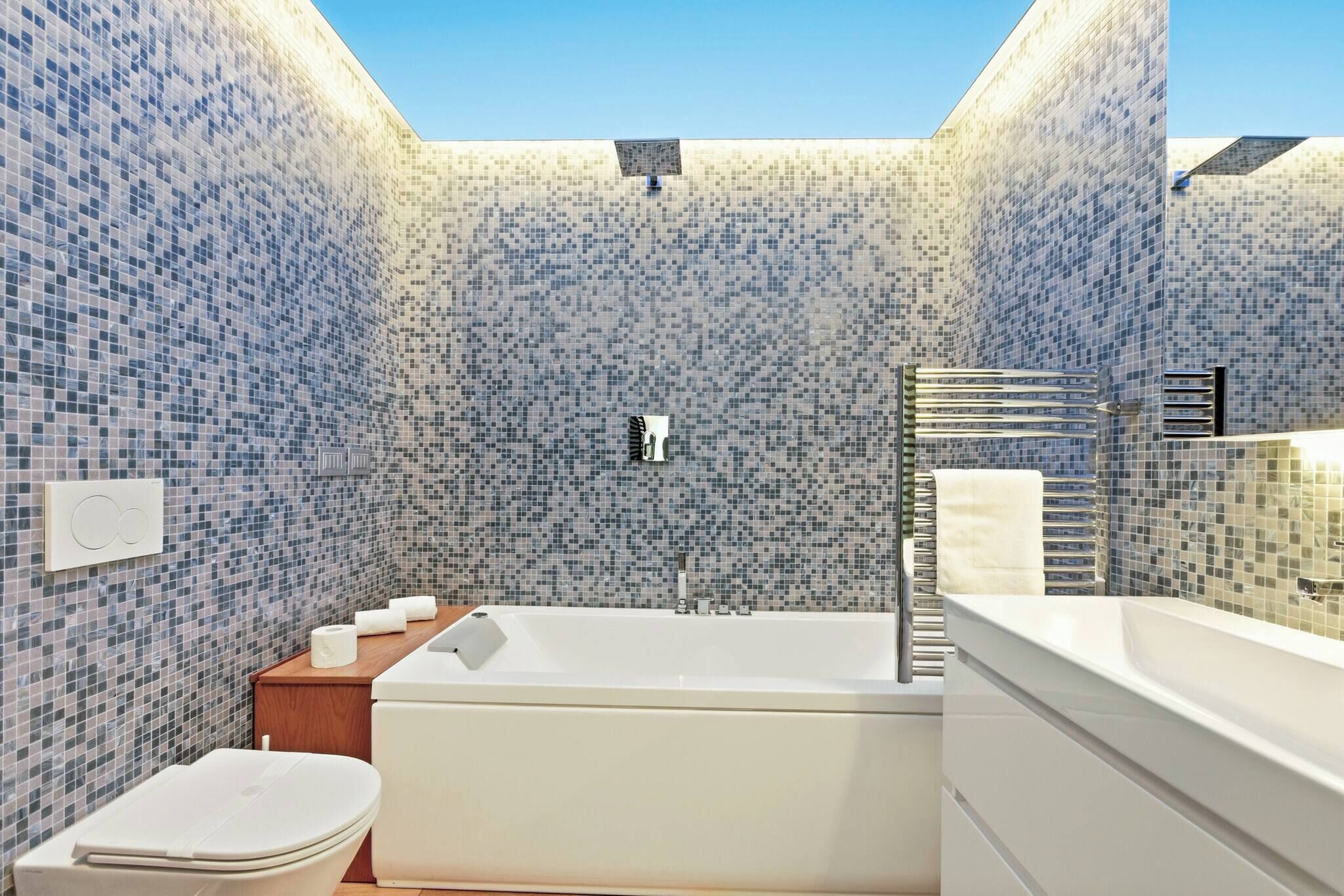 A modern bathroom with mosaic tile walls, a freestanding bathtub, a toilet, and a wooden vanity.