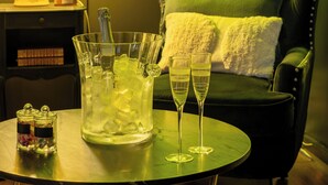 A green-lit room with a neon sign reading "Luxuria", a green upholstered chair, a table with two glasses of champagne, and a floral lamp. - Luxuria Love Room Avignon (Avignon)