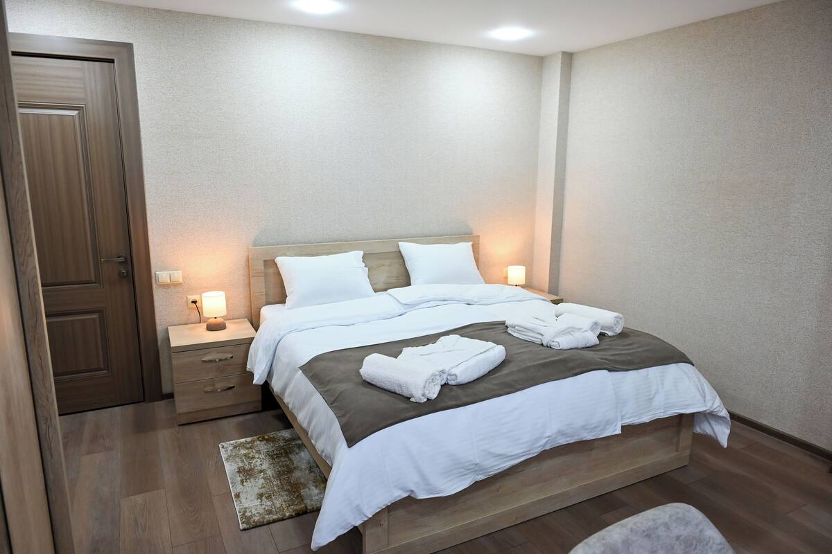 Una cama bien tendida con sábanas blancas y una manta marrón, una mesita de noche de madera con una lámpara, y una puerta de madera.
