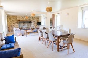 Een ruime woonkamer met een stenen openhaard, een eettafel met stoelen en een blauwe bank met kussens.