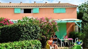 Une maison avec un toit en tuiles, des volets verts et un jardin où poussent des fleurs roses et des plantes à feuillage vert.