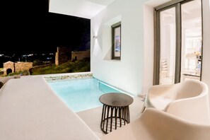 Un área de piscina moderna al aire libre con un muro de piedra, una pequeña mesa redonda y dos sillas blancas.