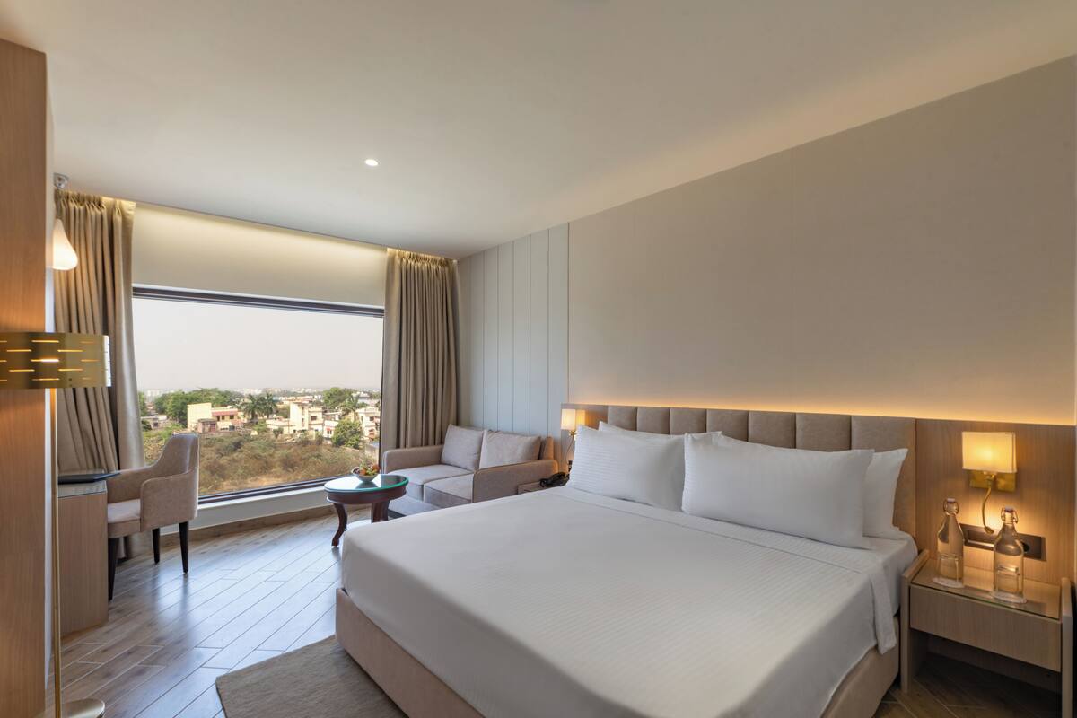 A hotel room with a large bed, a sitting area with a chair and a small table, a nightstand with a lamp, and a view of the city through the window.