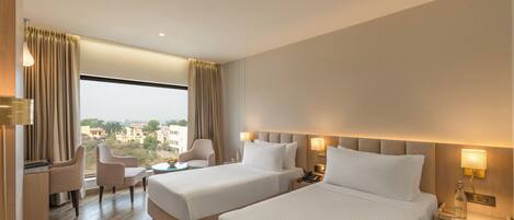 A hotel room with two beds, a desk, a chair, and a view of the city.