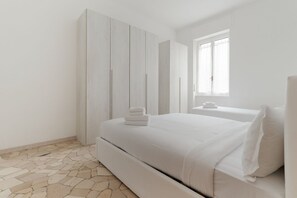 Um quarto com cama, mesa de cabeceira, guarda-roupa e janela.