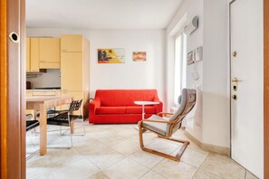 A room with a red sofa, a wooden rocking chair, a small white table, and a kitchen area with yellow cabinets. - Hostmate - Milano - Plinio - Città Studi (Milano)