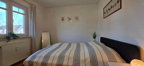 A bedroom with a bed, a radiator, a window with curtains, a bedside lamp, and wall art. - Vacation apartment E3 - "Moorfrosch" - 2-room apartment on the 1st floor near the beach (Graal-Müritz)