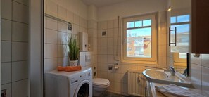 A bathroom with a washing machine, toilet, radiator, and a window with a view of buildings. - Vacation apartment E3 - "Moorfrosch" - 2-room apartment on the 1st floor near the beach (Graal-Müritz)