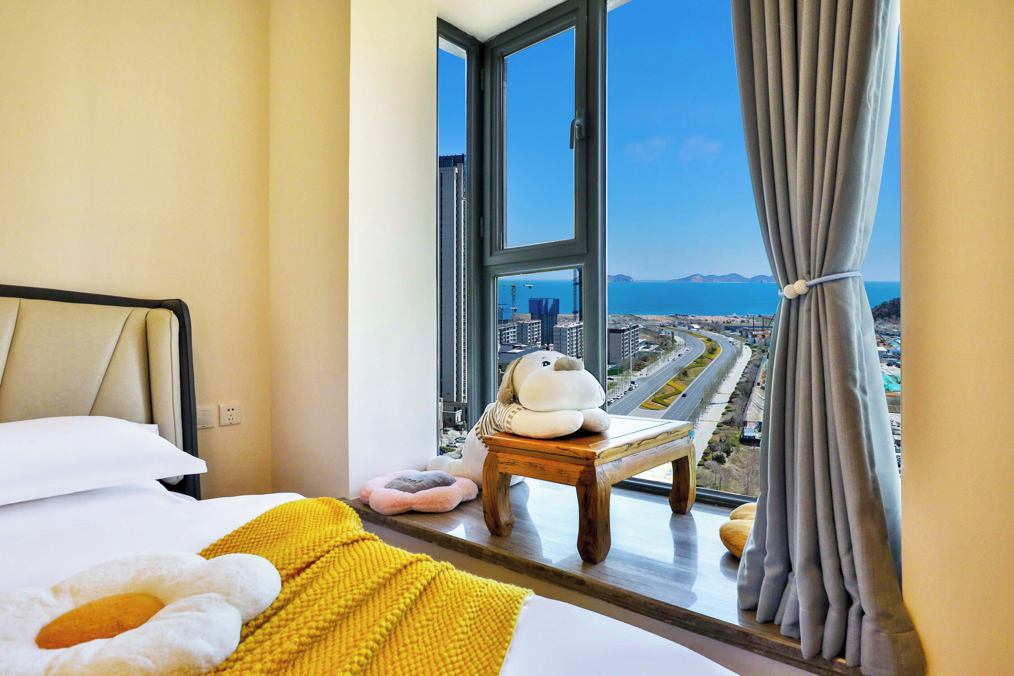 A hotel room with a large window, a bed with a yellow blanket, a small wooden stool, and a plush toy.
