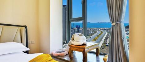 A hotel room with a large window, a bed with a yellow blanket, a small wooden stool, and a plush toy.