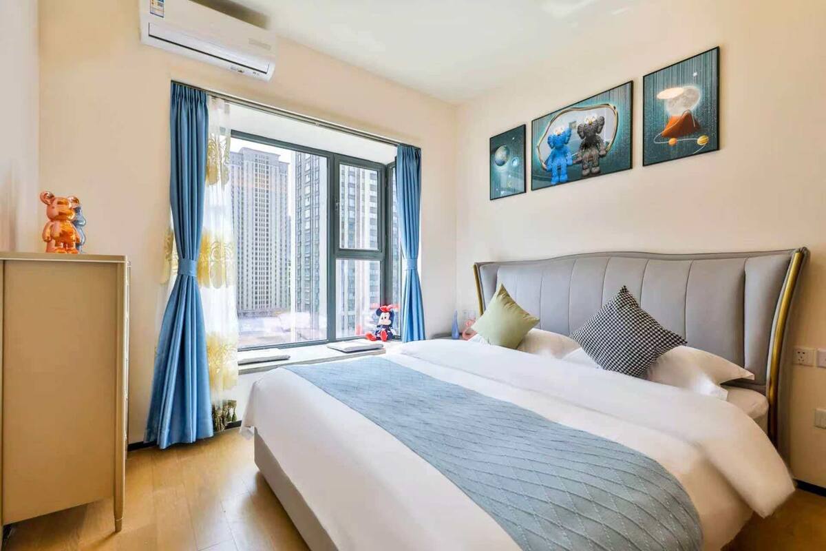 Ein Hotelzimmer mit einem großen Bett, einem Fenster mit Vorhängen, einer Wand-Klimaanlage und drei gerahmten Bildern.