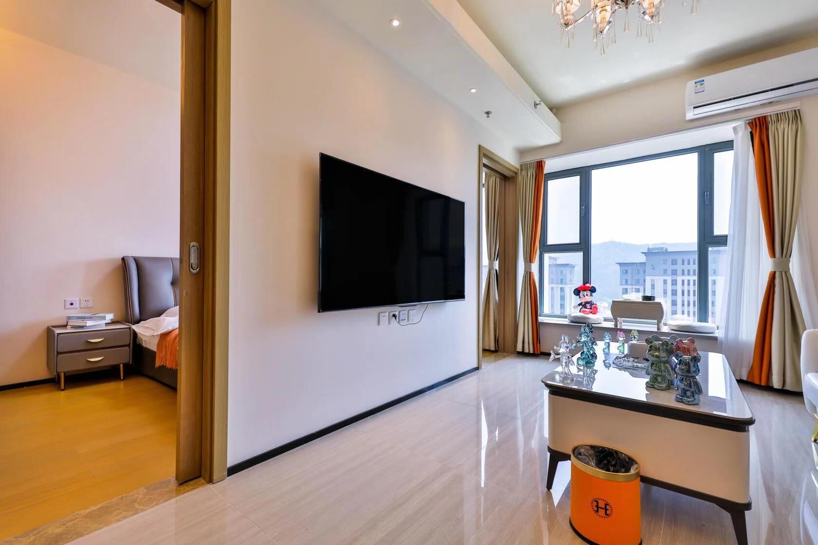 A modern hotel room with a flat-screen TV, a bedside table with a lamp, a window with curtains, and a view of buildings outside.