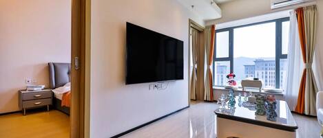 A modern hotel room with a flat-screen TV, a bedside table with a lamp, a window with curtains, and a view of buildings outside.