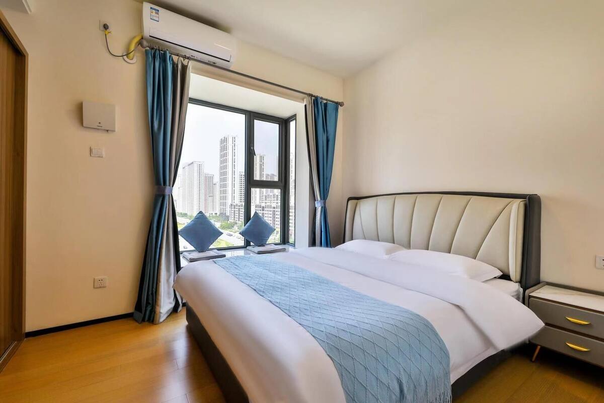 Ein modernes Hotelzimmer mit einem großen Bett, Blick auf hohe Gebäude und einer Klimaanlage an der Wand.
