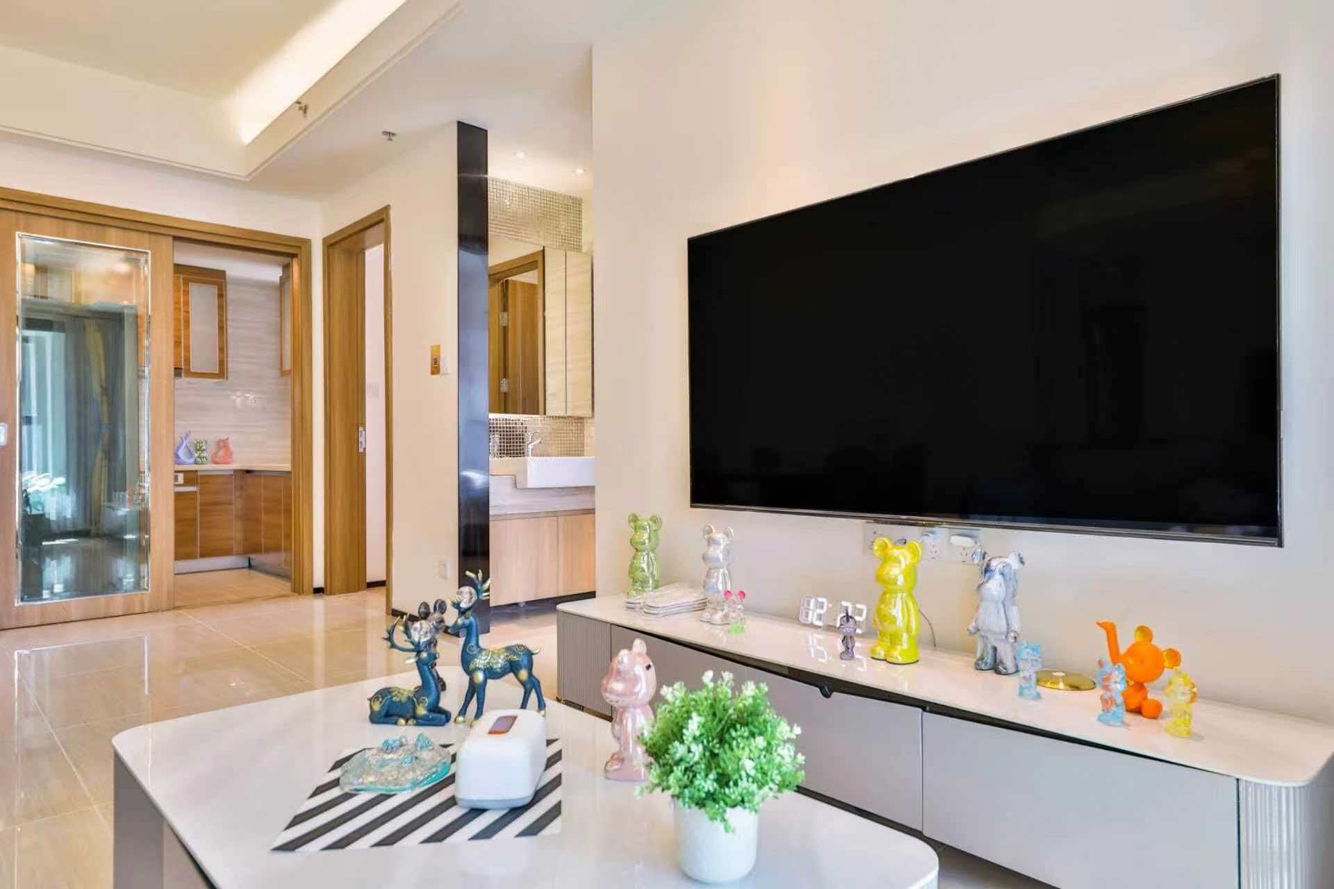 Un salon moderne doté d’un grand téléviseur à écran plat fixé au mur, d’une table basse agrémentée d’objets décoratifs et d’un espace cuisine visible à travers une porte ouverte.