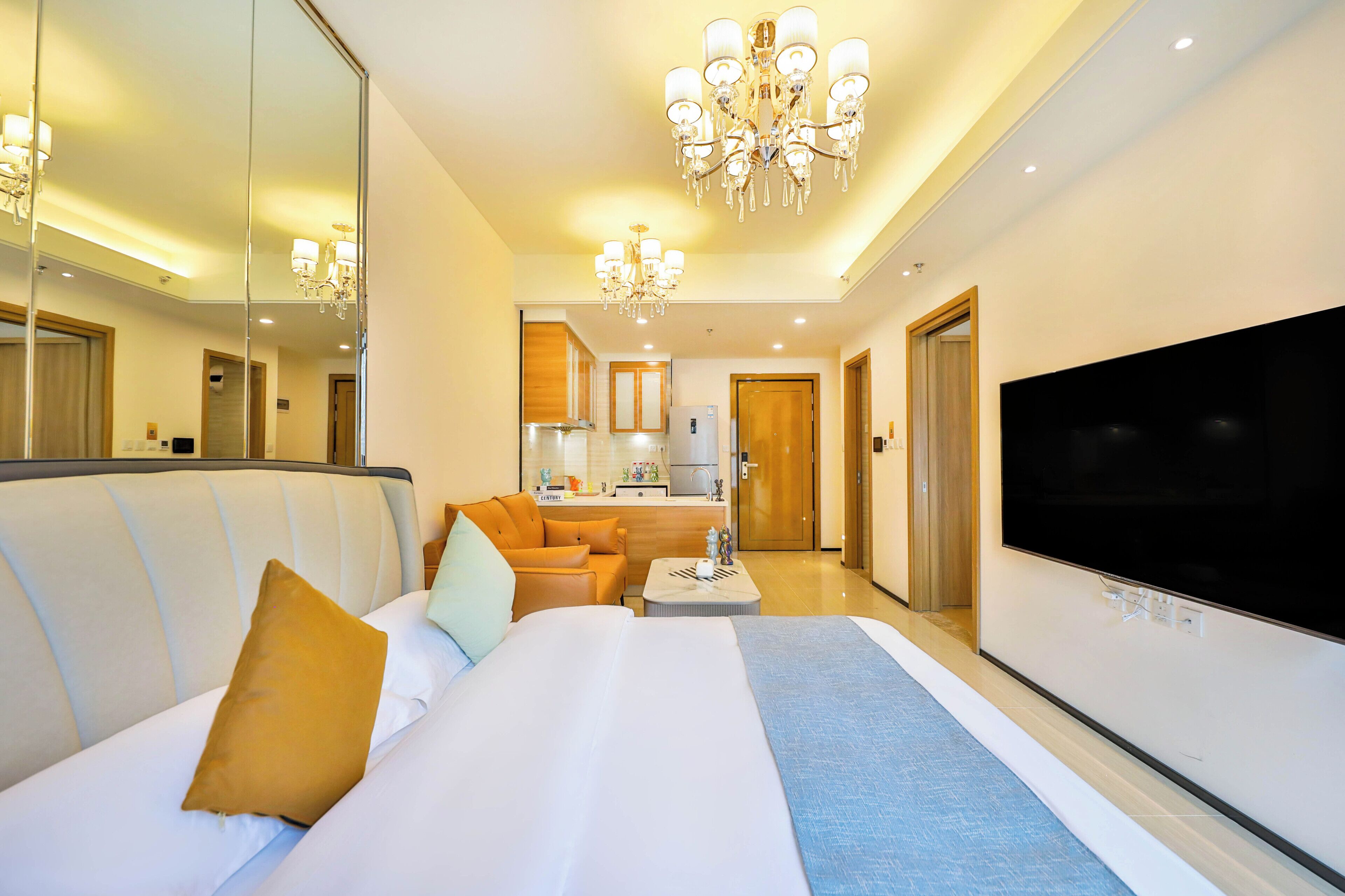 A hotel room with a large bed, a sofa, a TV, and a kitchen area in the background.