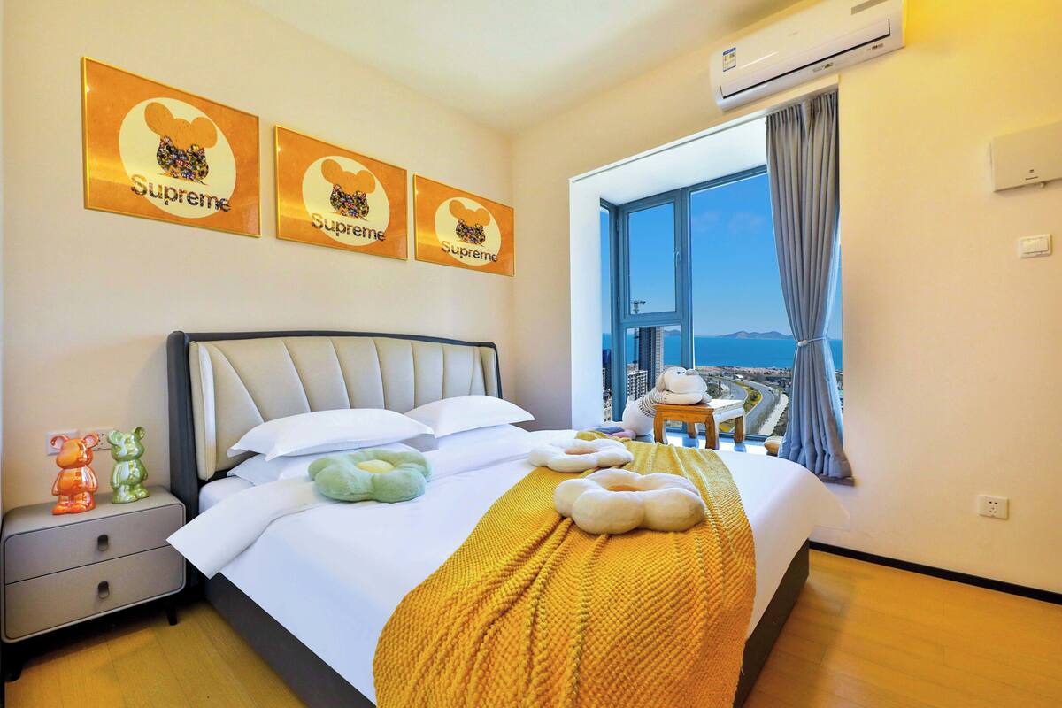 Ein Hotelzimmer mit Bett, Kissen, Decke, Nachttisch, Teddybär, Fenster mit Blick und drei Wandbildern der Marke 'Supreme'.