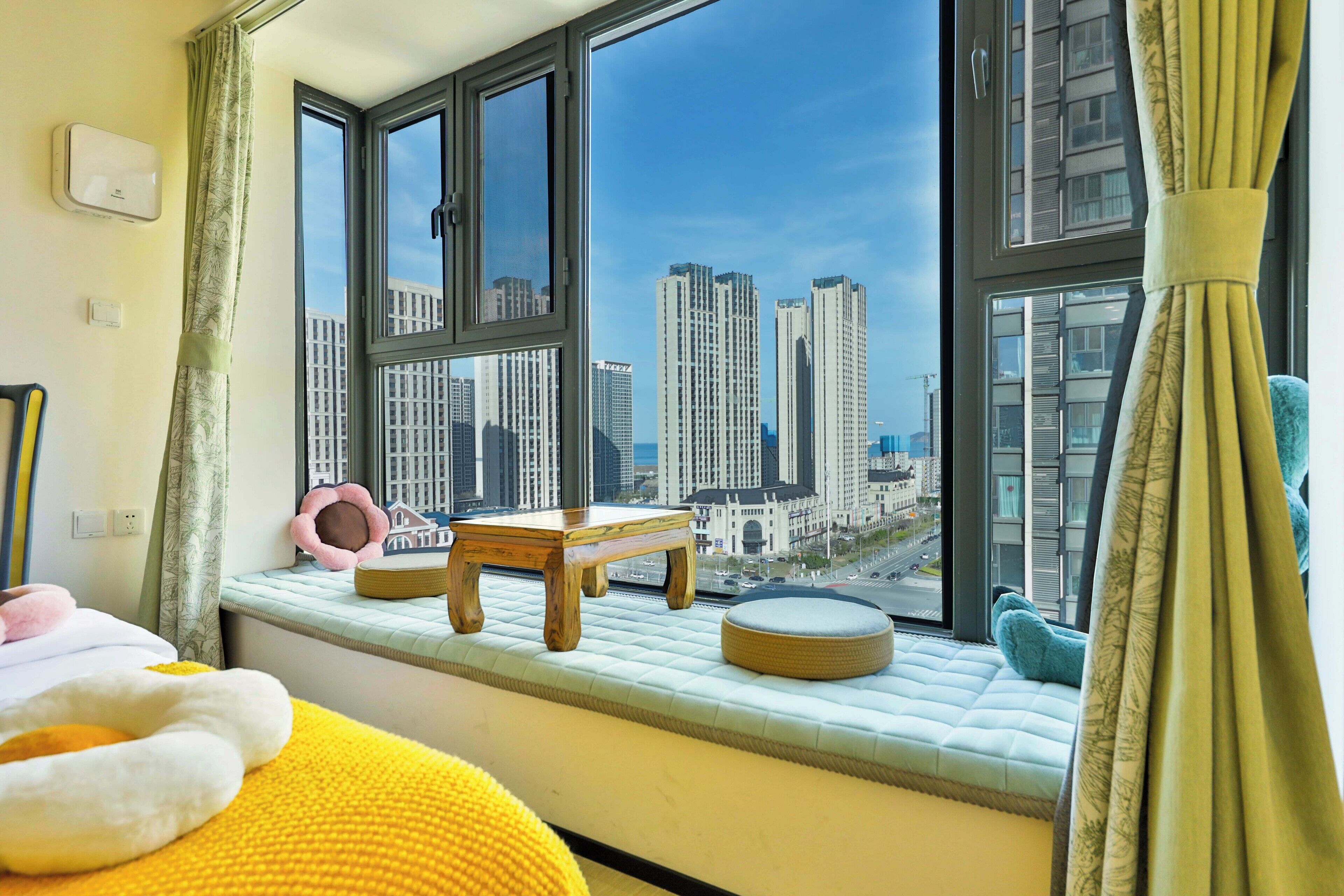 A bedroom with a large window, a bed, a small wooden table, and a cushioned bench.