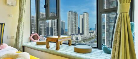A bedroom with a large window, a bed, a small wooden table, and a cushioned bench.