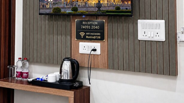 A television mounted on a wall displaying a building, a reception sign below it, and a power outlet with a plug. - The Manthan Heights (Vrindavan)