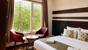 A hotel room with a bed, two chairs, a small table, and a window with curtains. - The Manthan Heights (Vrindavan)