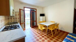 Una cocina compacta con estufa a gas, un lavamanos y un mueble pequeño. Hay un comedor con mesa y sillas, y un televisor sobre un soporte.