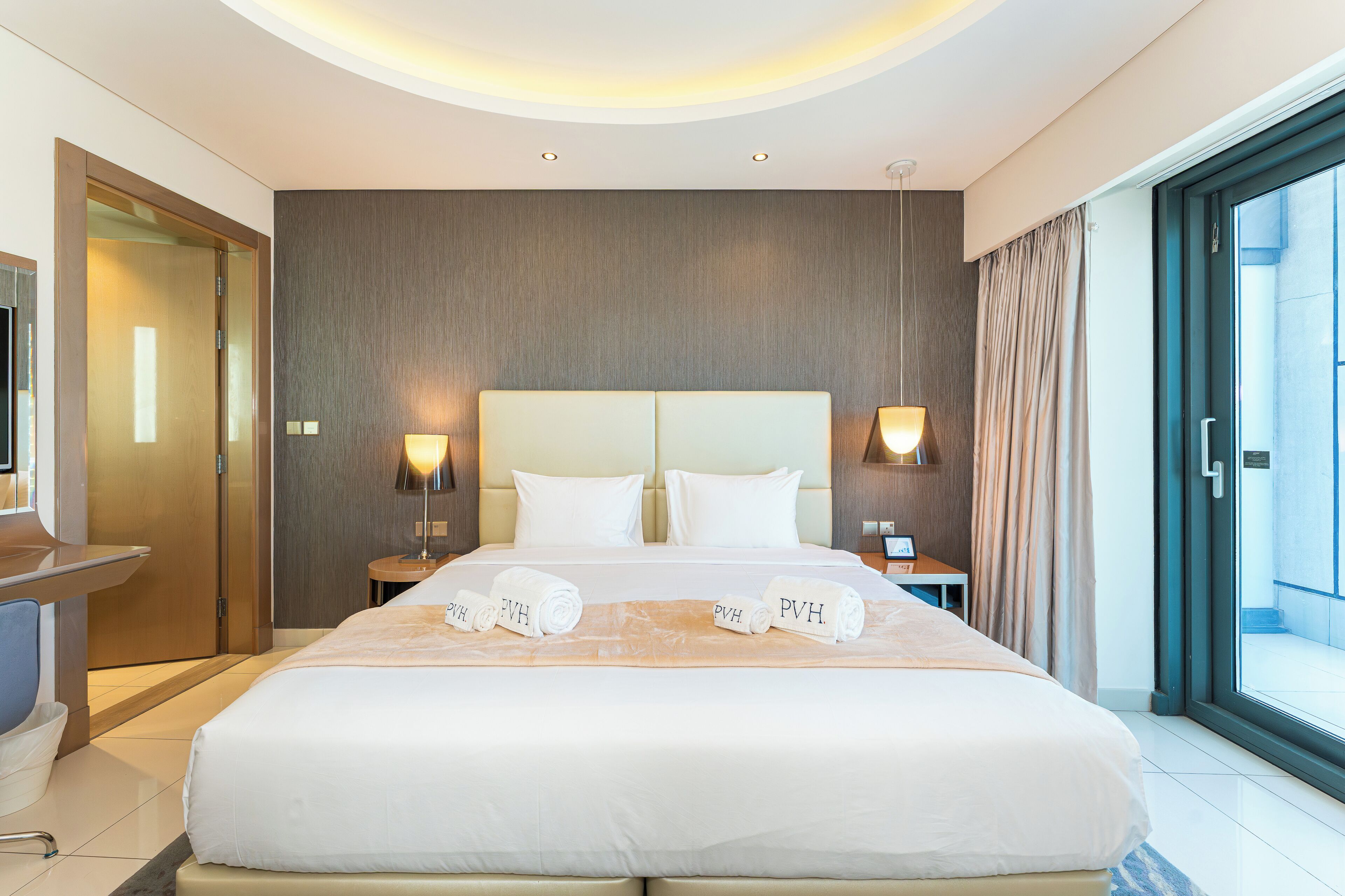 A modern hotel room with a large bed, two bedside tables with lamps, a desk, and a sliding glass door to a balcony.