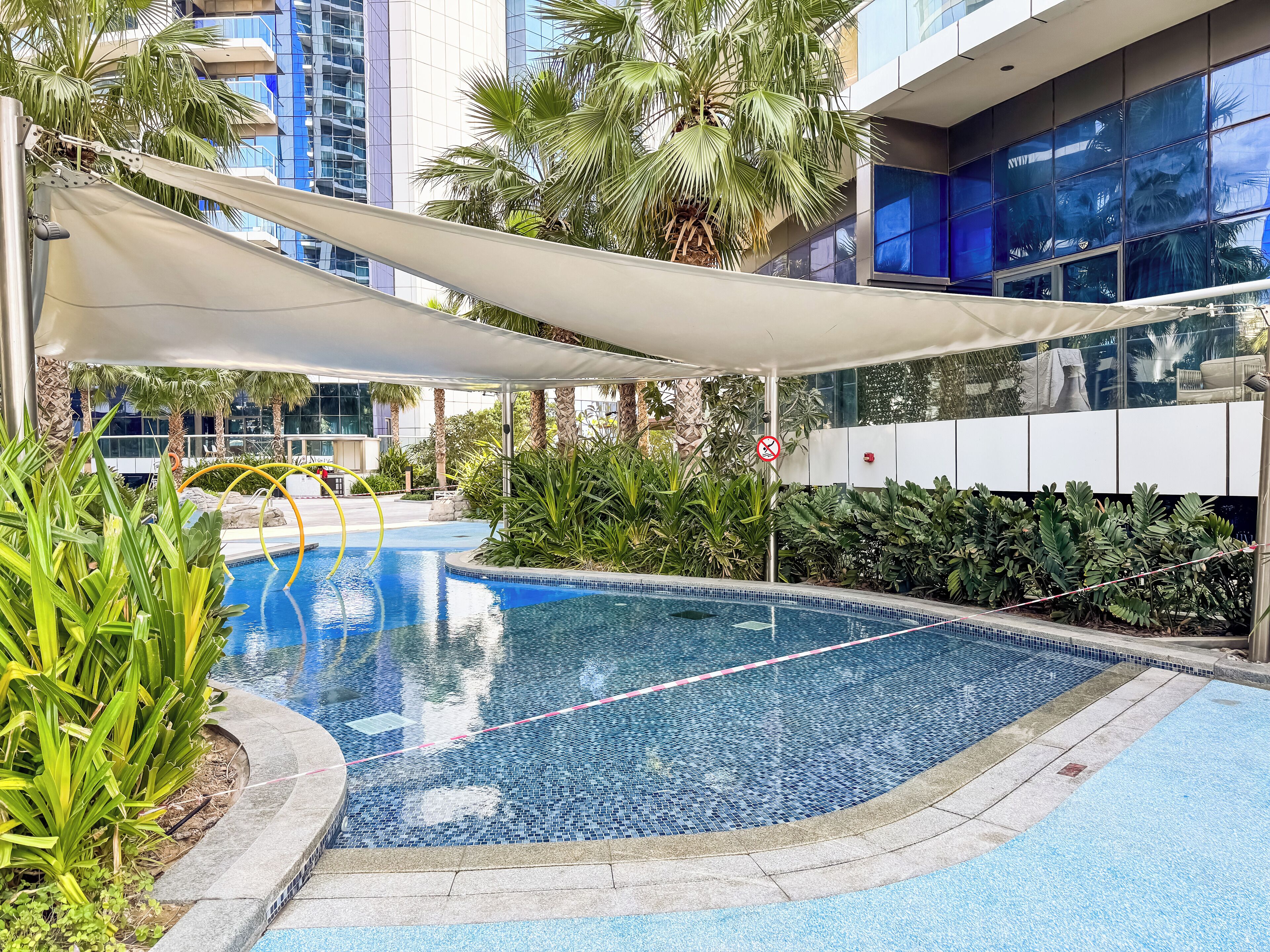 Une piscine dont le bord est revêtu de carrelage, entourée d’une végétation luxuriante et avec un bâtiment moderne en arrière-plan.