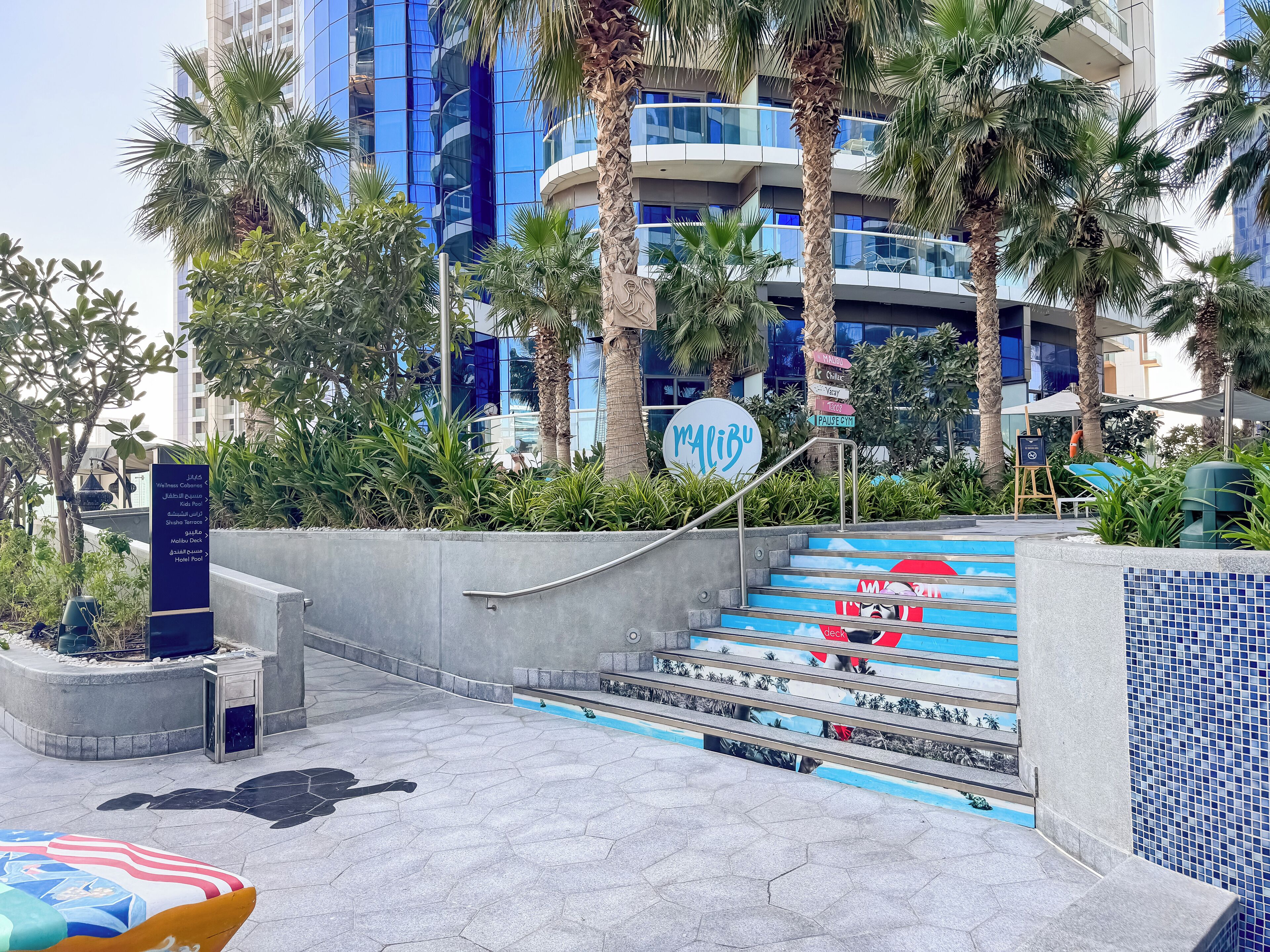 A hotel entrance with a palm tree-lined pathway, a sign for 'Malibu', and a staircase leading to a pool area.