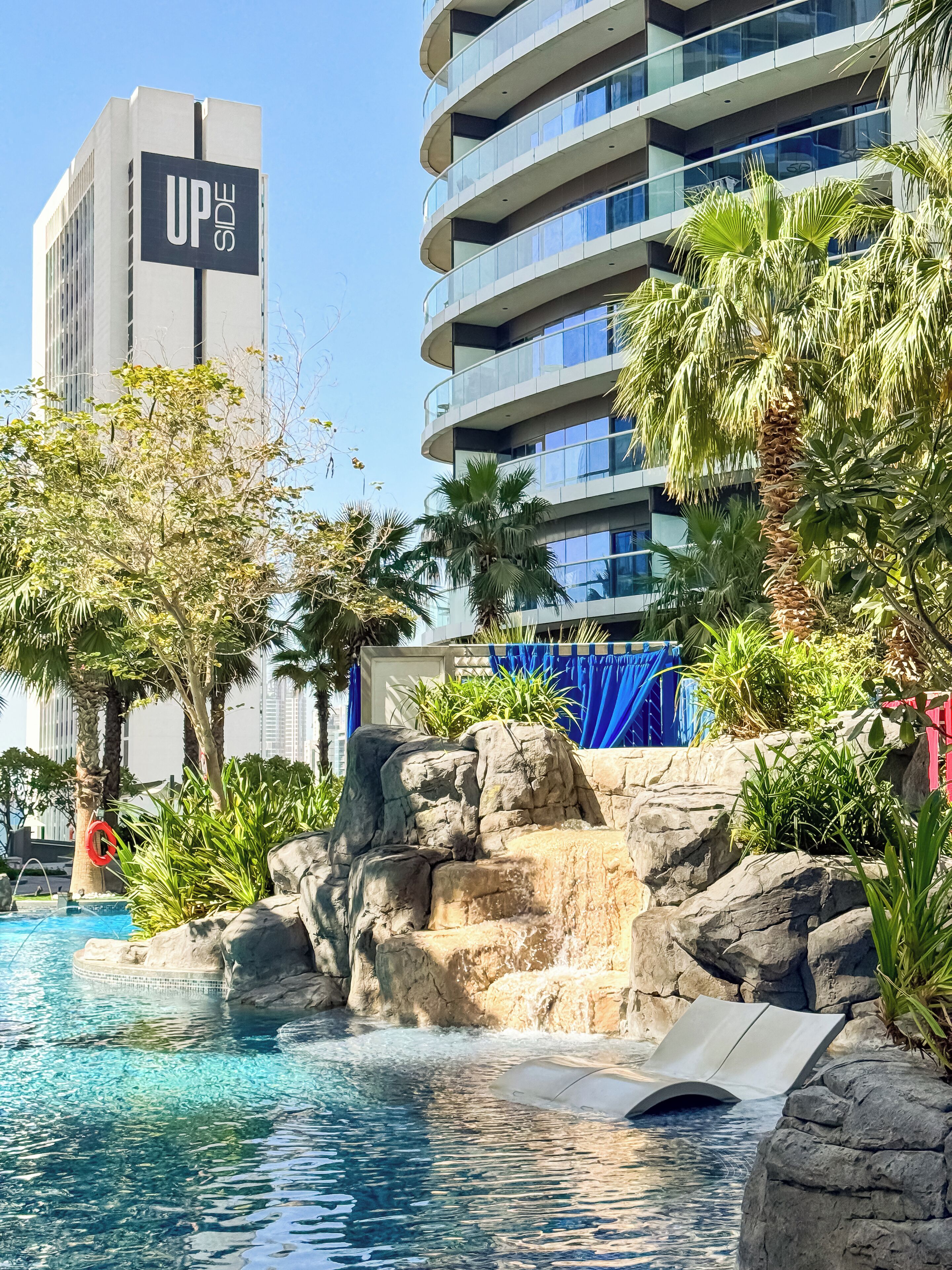 Una piscina con cascada, rodeada de rocas y palmeras, con un edificio moderno al fondo.