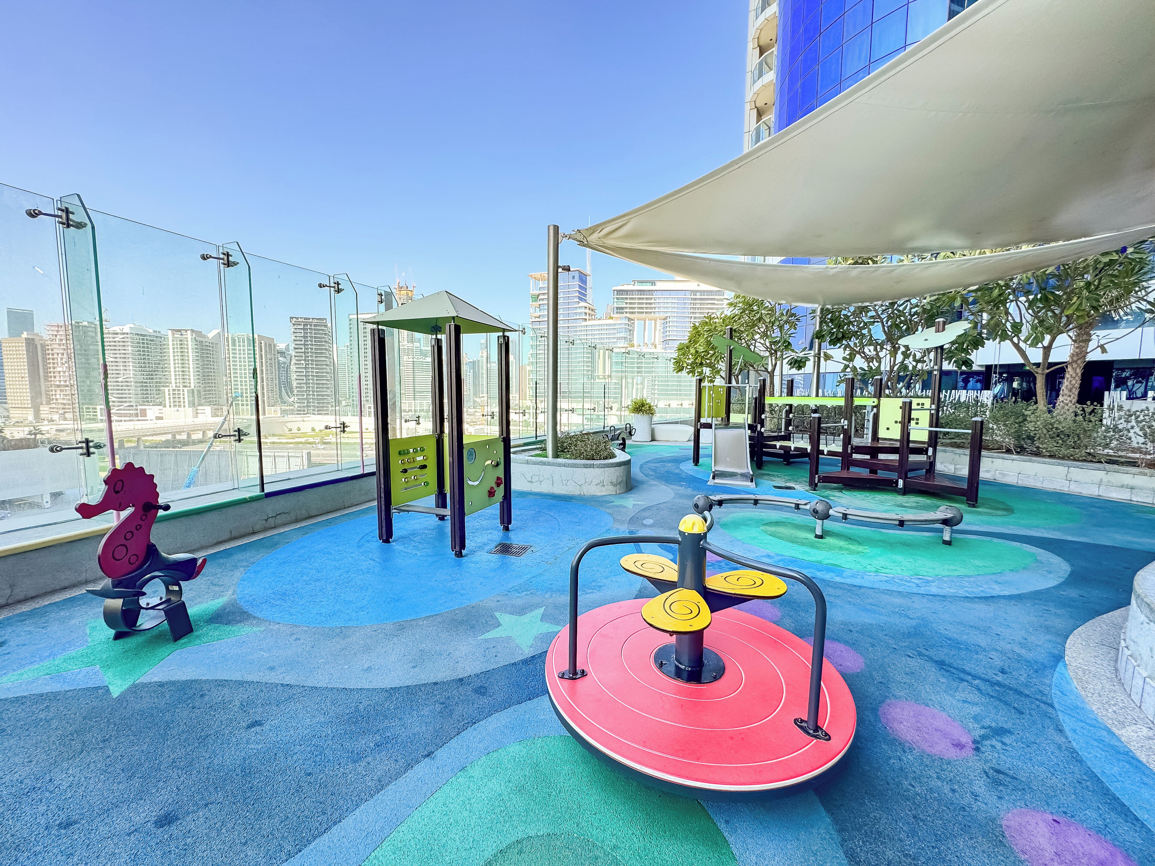 A rooftop playground with exercise equipment, a seahorse swing, and a view of the city skyline.