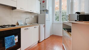Eine Küche mit weißen Schränken, einem Gasherd, einem Spülbecken, einem Kühlschrank und einem Fenster mit Blick auf Gebäude.