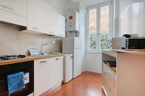 A kitchen with white cabinets, a gas stove, a sink, a refrigerator, and a window with a view of buildings. - San Pietro Comfy Apartment by Rentbeat (Roma)
