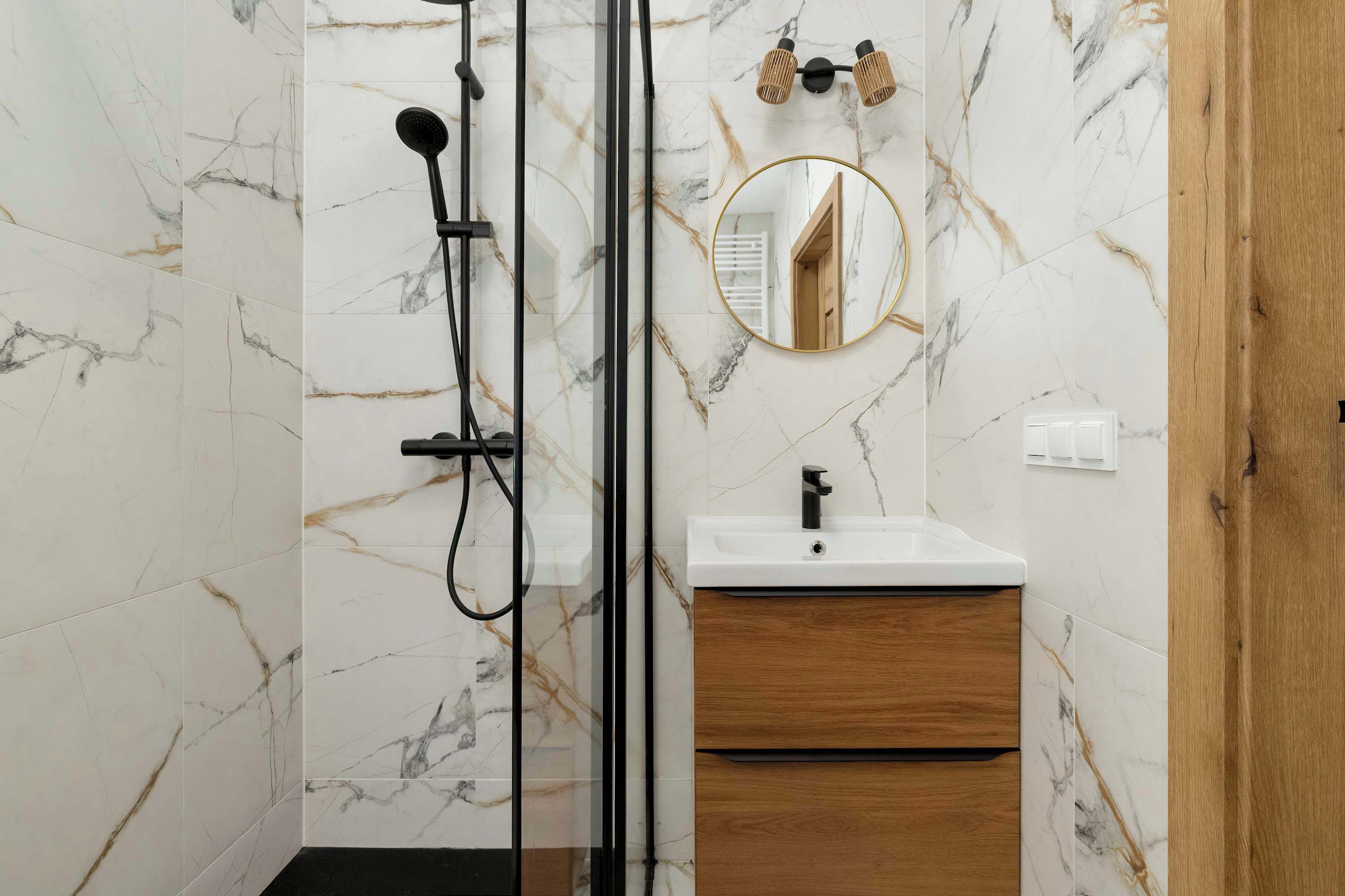 Un baño con cabina de ducha de vidrio, tocador de madera con lavabo blanco y pared con azulejos de mármol.