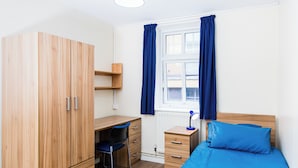 Une chambre avec une armoire en bois, un bureau, une chaise, un lit avec une literie bleue et une fenêtre avec des rideaux.