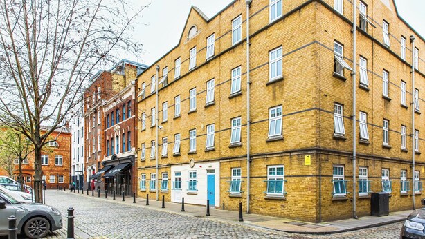 Un immeuble en briques de plusieurs étages, avec une rue pavée, une porte bleue et un arbre au premier plan.