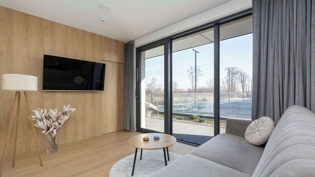 Una sala de estar moderna con un televisor de pantalla plana montado en una pared de madera, una mesa de centro redonda de madera con elementos decorativos, un sofá gris con un cojín blanco, y un amplio ventanal que ofrece vistas a un estacionamiento y árboles.