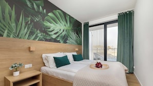 Una habitación de hotel con cama, mesita de noche con planta, mural en la pared y vistas por la ventana.