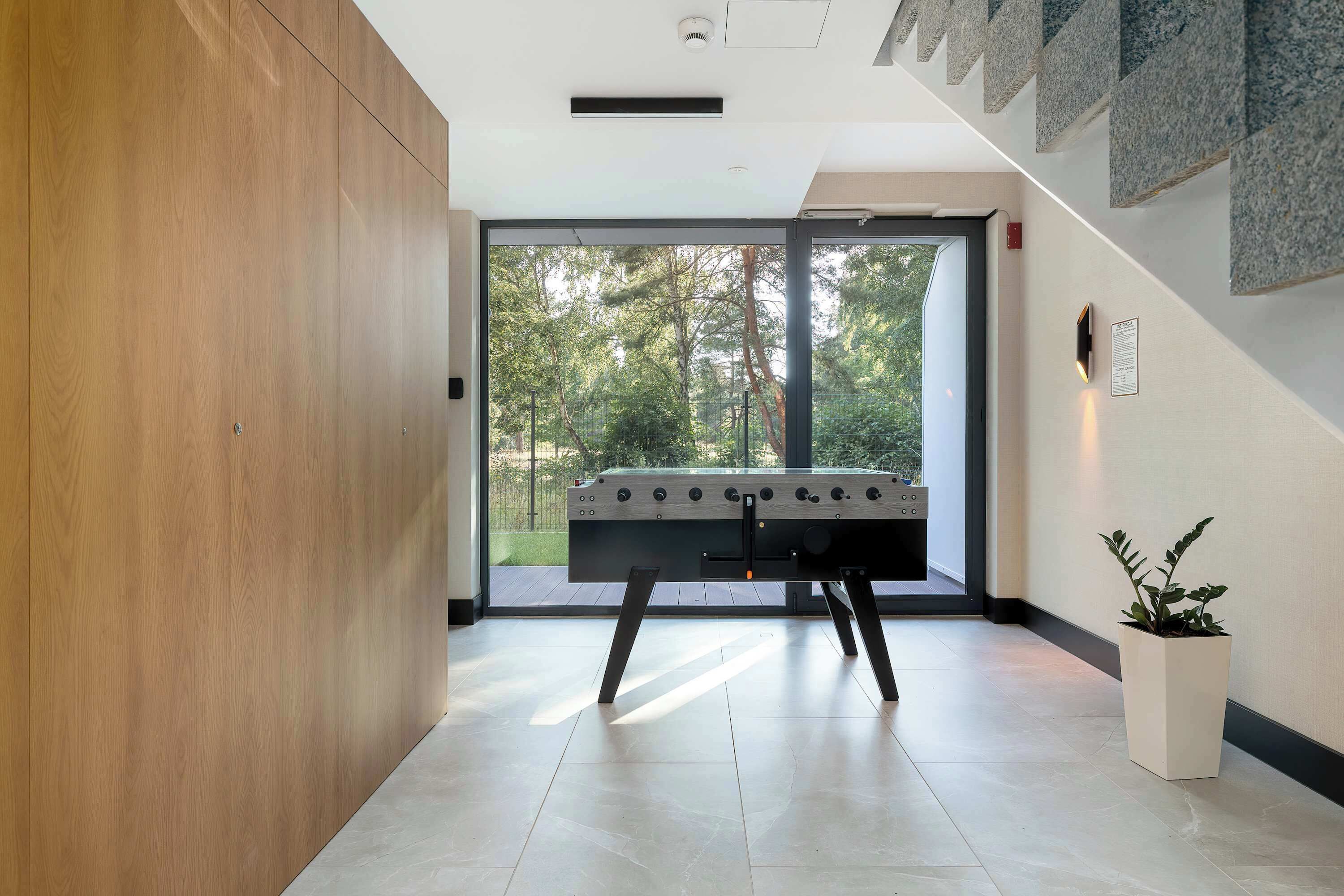 A modern indoor space with a foosball table, a wooden panel wall, a potted plant, and a view through a glass door.