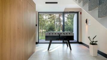 A modern indoor space with a foosball table, a wooden panel wall, a potted plant, and a view through a glass door.