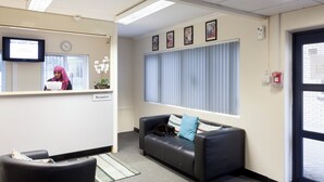 A reception area with a reception desk, a black leather sofa with a blue pillow, a clock on the wall, framed pictures, and a window with blinds. - Harrow Hall Northwick Park  (Harrow)