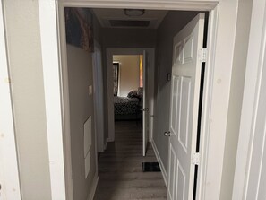 Ein schmaler Korridor mit weißen Wänden und Holzboden, der zu einem Schlafzimmer mit einem gemusterten Bettüberwurf führt.
