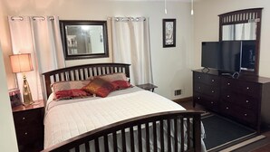 Una camera da letto con un letto, una cassettiera, una televisione, uno specchio, una ventola a soffitto e una finestra con tende.