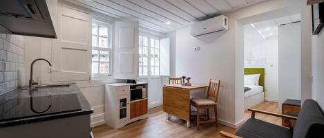 A compact kitchen with a black countertop, a microwave, and a small cabinet. There is a dining area with a wooden table and chairs, and a bed with a green headboard in the background.